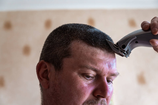 A middle-aged man cuts his hair. The person cuts the hair on his head with an electric hair clipper. Short brown hair. Inside a living room. Selective focus. - Photo, Image