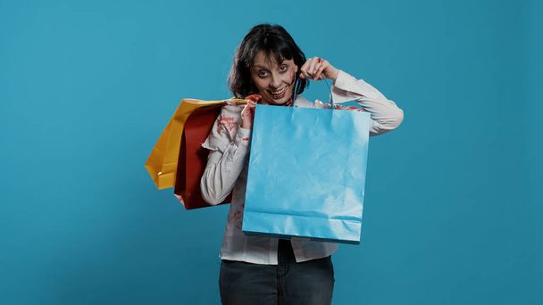 Sinistere angstaanjagende zombie met kleurrijke boodschappentassen van winkelcentrum op blauwe achtergrond. Gevaarlijk agressief uitziend hersenloos lopend lijk met bloederige wonden met kortingsaankopen.. - Foto, afbeelding