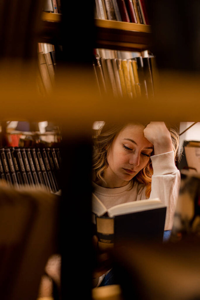 Kolejli kız kütüphanede oturmuş başını tutarak kitap okuyor. - Fotoğraf, Görsel