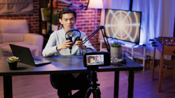 Asyalı adam kulaklık ürününü kamerada inceliyor, sosyal medyada podcast bölümü için video çekiyor. Kulaklık önermek için canlı yayın vlog 'u, Web istasyonunda prodüksiyonu göster. - Fotoğraf, Görsel