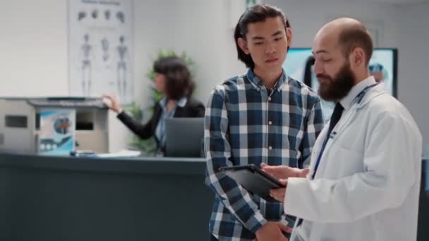 Hausarzt im Gespräch mit asiatischem Mann über Diagnose auf digitalem Tablet, mit Gesundheitsbericht zur Untersuchung im Wartebereich eines Krankenhauses. Diverse Menschen plaudern in Empfangshalle. - Filmmaterial, Video