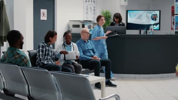 Африканский врач-американец разговаривает с азиатским пациентом в зоне ожидания, проводит консультации в приемном холле больницы с медицинским персоналом. Различные люди в чате в комнате ожидания. - Кадры, видео