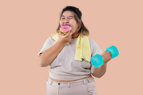 Ασιατική μεγάλο κορίτσι κάνει ασκήσεις φυσικής κατάστασης με αλτήρες και τρώει γλυκό ντόνατ μαζί απομονωμένο. - Φωτογραφία, εικόνα