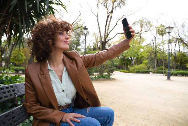 Красива і сексуальна жінка з кучерявим волоссям фотографує себе зі своїм мобільним телефоном, сидячи в парку. Жінка щаслива під час фотографування. Концепція мобільного телефону
 - Фото, зображення