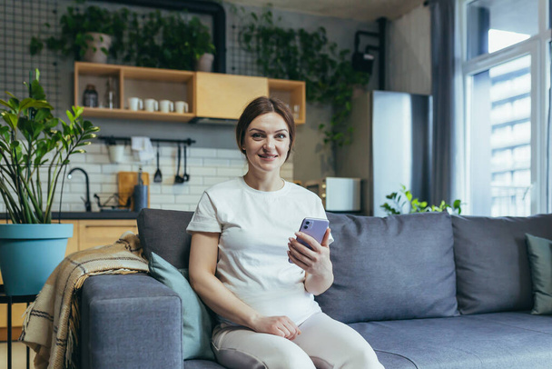 Happy pregnant woman at home rejoices with phone smiling at home and looking at camera - Foto, Bild