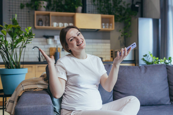 Happy pregnant woman at home rejoices with phone smiling at home and looking at camera - Фото, зображення