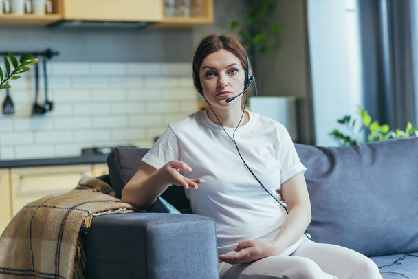 Pregnant woman working remotely at home, using video call headset, consultant sitting on sofa looking at webcam and talking - Foto, Imagen