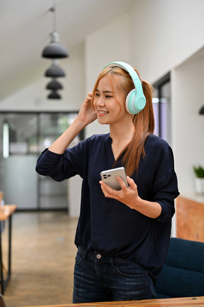 Retrato de una joven asiática alegre con unos audífonos azules de moda sosteniendo un smartphone, escuchando la música. Concepto de entretenimiento musical - Foto, imagen