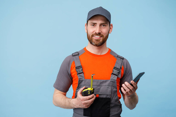 Homem construtor em conversa uniforme no telefone celular e segurando uma fita métrica no fundo do estúdio azul.  - Foto, Imagem