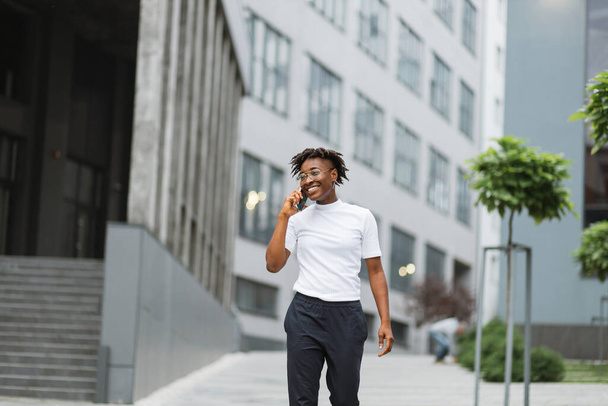 Photo of amazing pretty african american lady, spending time walking on city street after university lectures, holding telephone while speaking with best friend. - Фото, изображение