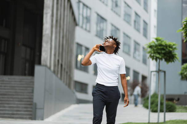 Photo of amazing pretty african american lady, spending time walking on city street after university lectures, holding telephone while speaking with best friend. - Foto, immagini
