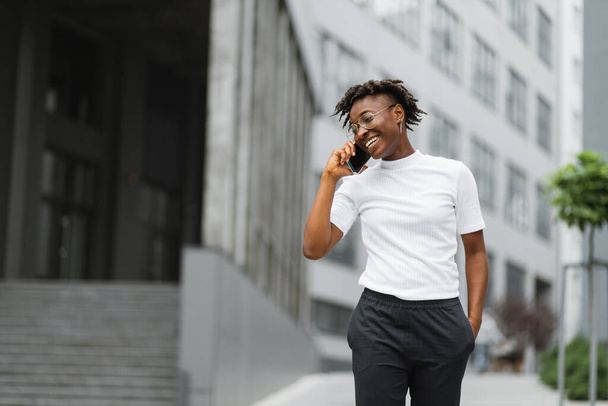 Portrait latéral d'une jeune femme afro-américaine souriante marchant et parlant avec un téléphone portable à l'extérieur. Gros plan portrait de jeune femme séduisante souriant et parlant sur smartphone. - Photo, image