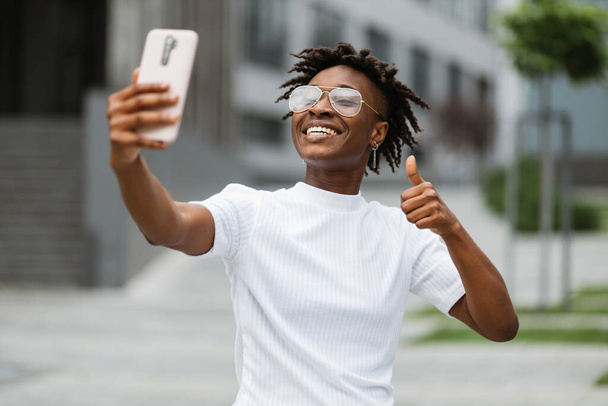 Young african-american woman girl student freelancer talking on phone having conversation with friends colleagues outdoors in modern city. Beautiful female taking selfie showing thumb up. - Foto, Imagem
