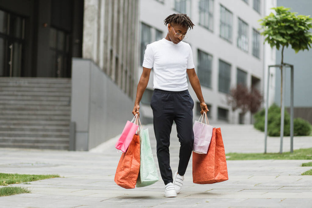 Pretty young African curly woman, wearing black pants and white t-shirt, walking at the street with colorful shopping bags, smiling and looking outside. Shopping in the city concept. - Foto, Imagen
