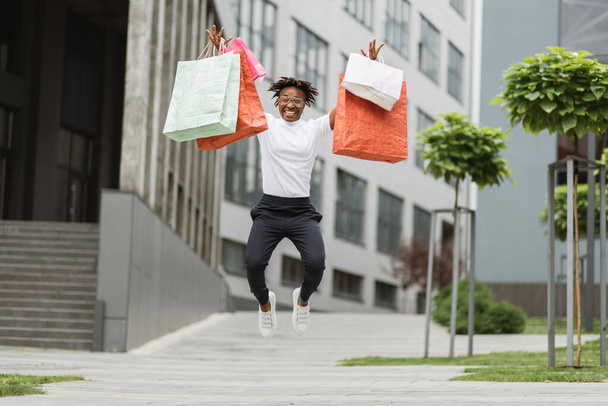 Beautiful happy African woman wearing black pants and stylish white t-shirt, posing outdoors in beautiful modern city with many colorful shopping bags while jumping with happiness new purchases. - Foto, Bild