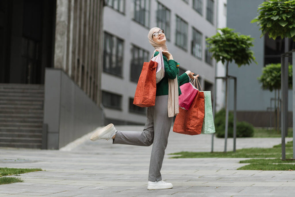 Pretty young muslim woman, wearing grey pants and green jacket, walking at the street with colorful shopping bags, smiling and looking outside. Shopping in the city concept. - Φωτογραφία, εικόνα
