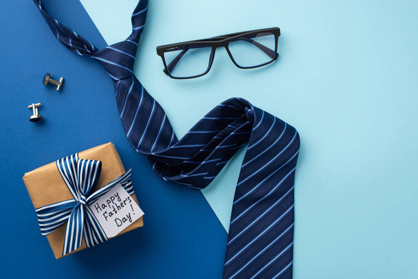 Father's Day concept. Top view photo of craft paper giftbox with silk ribbon bow and postcard glasses blue tie and cufflinks on bicolor blue background - Photo, Image