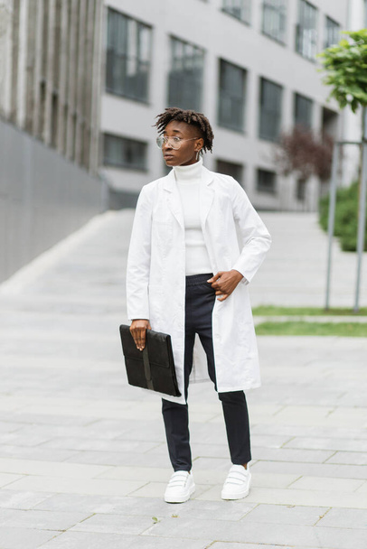 Portrait of young smart focused african female medical intern doctor with folder clipboard, standing outside modern hospital building. Medicine, education and healthcare concept. - Foto, Bild