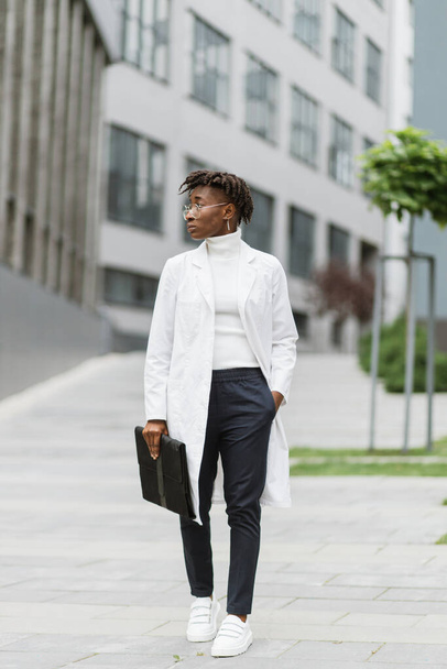 Portrait of young smart focused african female medical intern doctor with folder clipboard, standing outside modern hospital building. Medicine, education and healthcare concept. - 写真・画像