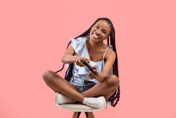 Full length of happy female gamer with joystick playing video game on chair over pink studio background. Joyful young black woman with controller taking part in computer gaming competition - Фото, изображение