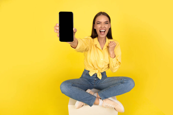 Yes. Emotional Woman Showing Cellphone With Empty Screen Shouting And Shaking Fist Advertising Mobile Offer Or Reading Great News Sitting On Yellow Studio Background. Technology And Gadgets. Mockup - Fotografie, Obrázek