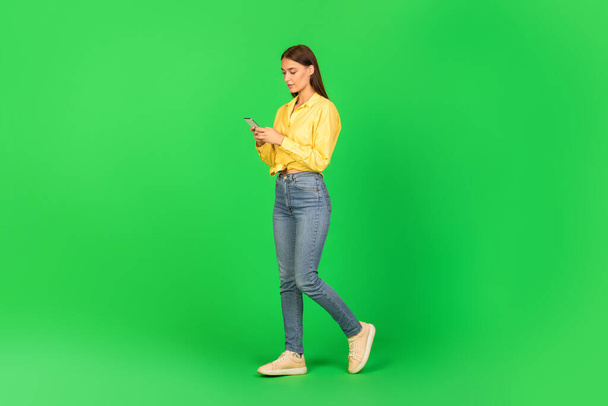 Mujer joven usando el teléfono móvil navegar por Internet y mensajes de texto caminando en el estudio sobre fondo verde. Comunicación móvil, nuevo anuncio de aplicación. Disparo de longitud completa - Foto, Imagen