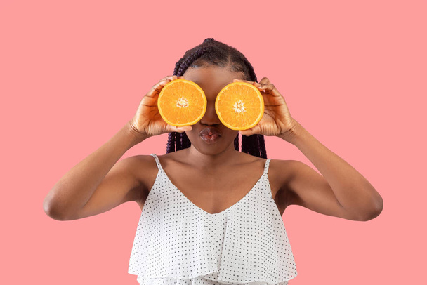 Summer detox. Millennial black woman with braids holding orange halves near her eyes, puckering lips for kiss on pink studio background. Young African American female holding citrus fruits near face - Foto, immagini