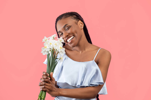 Vidám Y-generációs fekete hölgy kezében csokor nárcisz rózsaszín stúdió háttér. Szép millenniumi afro-amerikai nő egy csomó nárciszvirág ünneplő ünnep - Fotó, kép