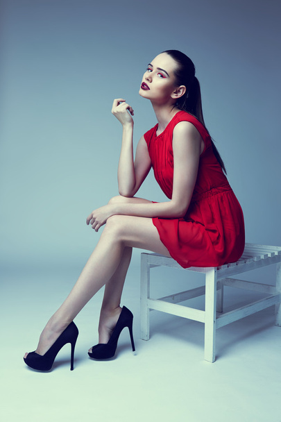 young elegant woman in red dress sit on stool, studio shot  - 写真・画像