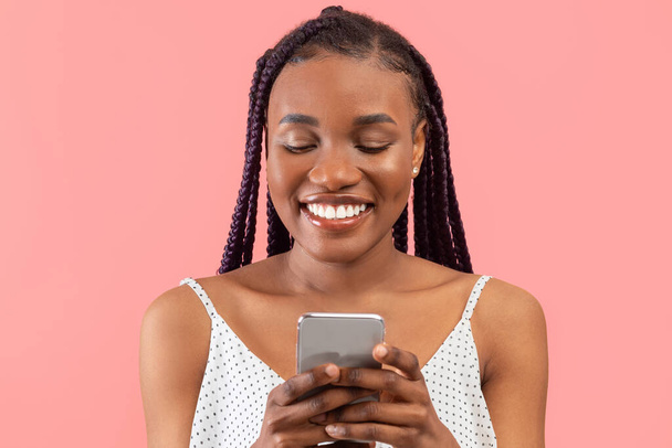 Cheerful millennial black woman using mobile phone, chatting on web, working or studying online over pink studio background. Pretty young African American lady watching video on smartphone - Foto, afbeelding