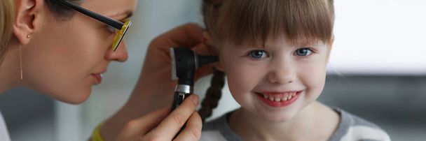 Porträt eines kleinen Mädchens, das lächelt, während der Arzt die Ohren mit einem speziellen Instrument untersucht. Ruhiges Kind, keine Angst vor Ärzten und Krankenhäusern. Vorsorgeuntersuchung, Kinderarztkonzept - Foto, Bild