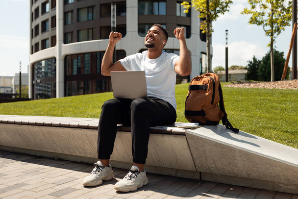 感情的なアフリカ系アメリカ人の男性は、ラップトップコンピュータで公園に座って、喜びで手を上げ、拳を振る、良いニュースを受け、昇進を得た、完成したプロジェクト - 写真・画像
