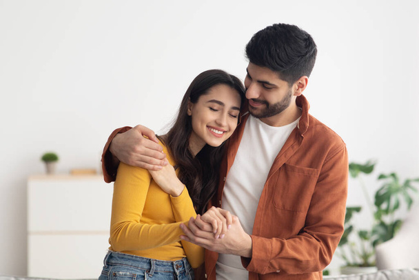 Loving Arabic Spouses Hugging Enjoying Time Together Standing Indoor. Middle Eastern Husband And Wife Embracing At Home. Happy Marriage And Romantic Relationship Concept - Photo, image
