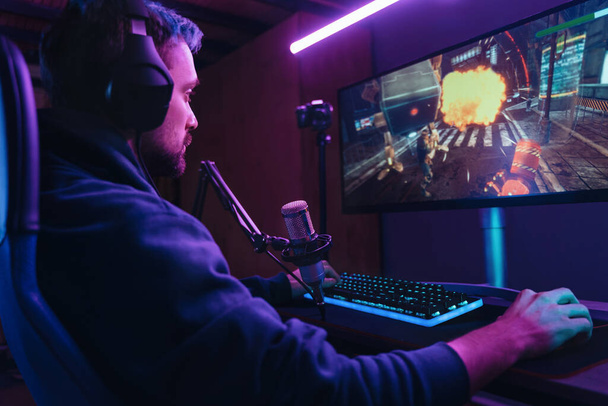 Sivunäkymä pro streamer pelaa videopeli kilpailu käyttää ammattimaista pelaamista setup. Pelaaja istuu pelituolilla käyttäen ammattimaisia kuulokkeita ja mikrofonia myöhään illalla kotona. Kyberurheilu - Valokuva, kuva