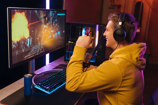 Side view of young caucasian pro gamer celebrating his victory in online video shooter game tournament. Exited cyber sportsman playing shooters on powerful pc at home at night, wearing headphones - Fotografie, Obrázek