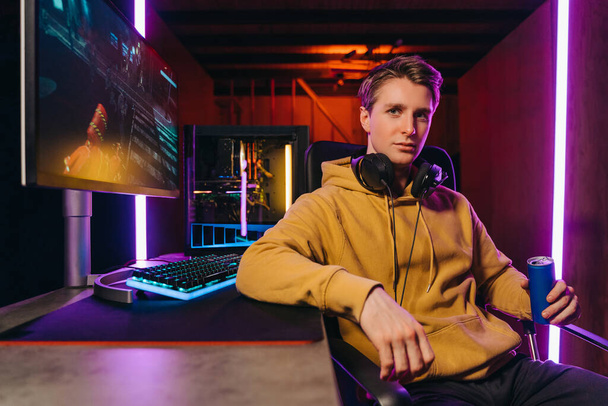 Portrait of young serious handsome pro gamer sitting at desk with gaming equipment, holding energy drink, using powerful PC for playing online video shooter games. Neon Colored Room, e-Sport concept  - Zdjęcie, obraz