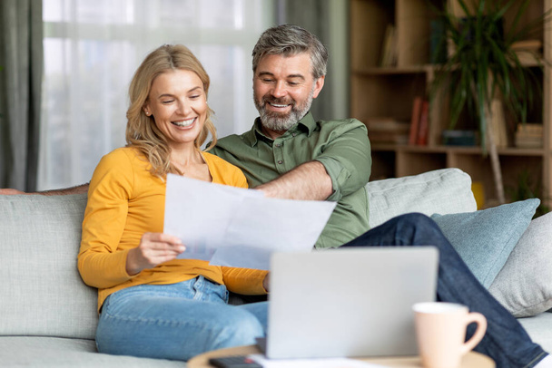 Концепция страхования семьи. Счастливая пара средних лет пара документов для чтения дома, улыбающиеся пожилые супруги проверяют финансовые документы, сидя вместе на диване в гостиной, бесплатное пространство - Фото, изображение