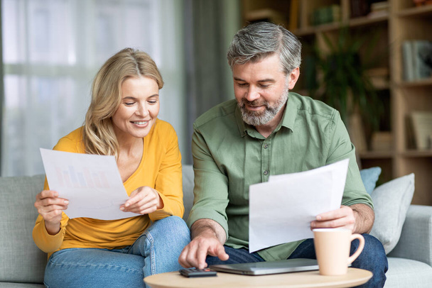 Glückliche Ehepartner mittleren Alters, die finanzielle Unterlagen prüfen und Ausgaben berechnen, während sie zu Hause gemeinsam das Familienbudget planen, lächelnde reife Ehepartner, die Zeitungen lesen und Taschenrechner benutzen. - Foto, Bild