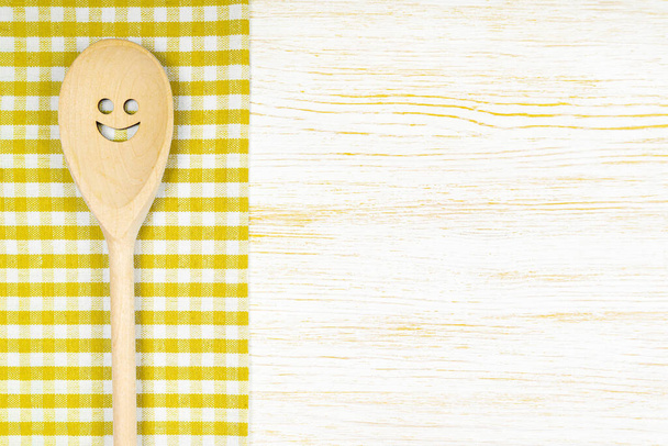 Cucchiaio in legno con sorriso su tovaglia a quadretti gialli su superficie in legno bianco. Mockup per menu o ricetta, ristorante, sito web con cucina. Cucina cibo sfondo, modello, piatto laici con spazio copia - Foto, immagini