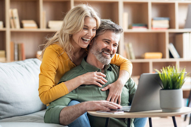 Sorrindo Cônjuges de meia-idade usando laptop juntos na sala de estar, feliz esposa madura e marido reserva férias on-line, verificando as ofertas de hotéis na Internet enquanto relaxa em casa, espaço livre - Foto, Imagem