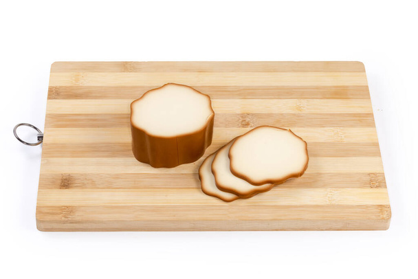 Trozo parcialmente cortado de queso ahumado procesado con película exterior de color marrón amarillento tradicional en la tabla de cortar de madera sobre un fondo blanco - Foto, Imagen