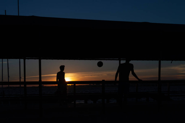 sylwetka dwojga młodych ludzi grających w piłkę nożną na chodniku portowym na plaży w Portugalii o zachodzie słońca - Zdjęcie, obraz