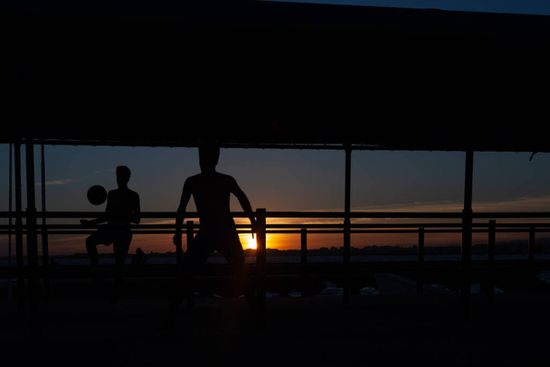 sylwetka dwojga młodych ludzi grających w piłkę nożną na chodniku portowym na plaży w Portugalii o zachodzie słońca - Zdjęcie, obraz