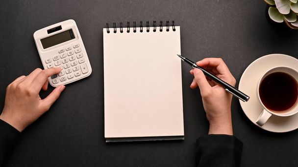 女性会計士は彼女のオフィスの机で財政収入と費用を計算する。トップ表示,女性を使用して計算機と空のスパイラルノートパッド上のノートを取る黒のテーブルの背景. - 写真・画像