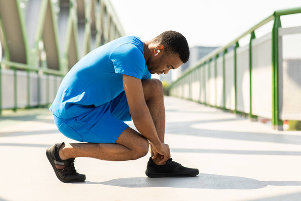 African guy in a blue sports suit tying his shoelaces on his sneakers before jogging. High quality photo - Фото, изображение