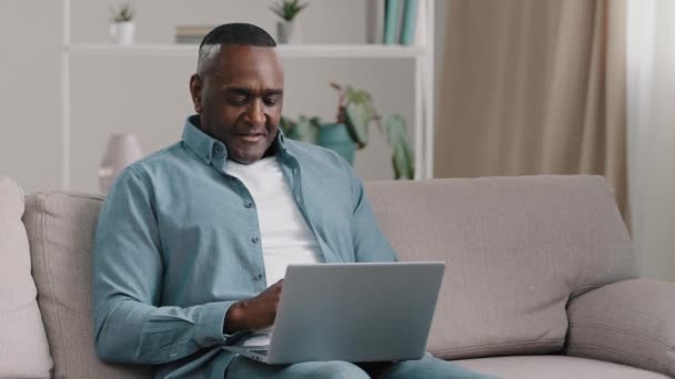 Dorosły Afroamerykanin siedzi na kanapie pisząc na laptopie przeglądając e-mail czytając dobre wiadomości wygrywając grę komputerową hazard wygrana loteria radując się zwycięstwem pokazując kciuk w górę dobry wynik uśmiechnięty - Materiał filmowy, wideo