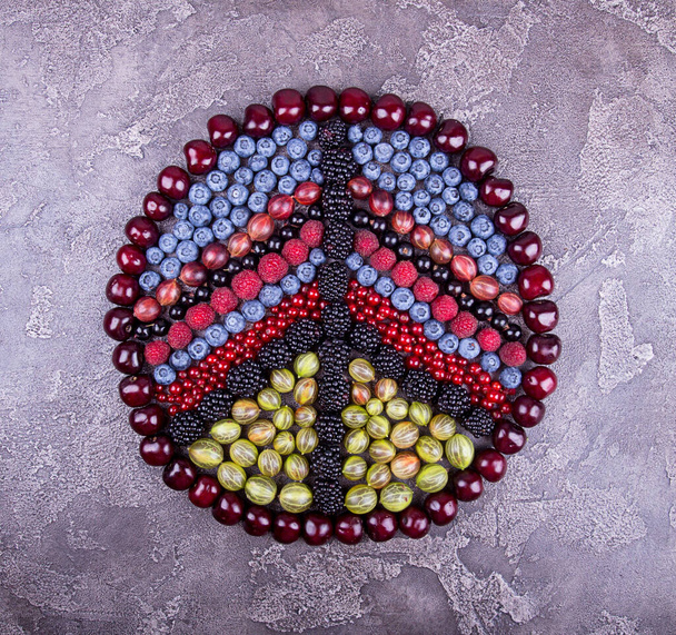 Sign pacifism of fruits and berries. Geometric pattern of summer fruits and berries: gooseberries, raspberry, red and black currant, blueberry, blackberries,  sweet cherry over on grey concrete background. Top view. - Photo, image