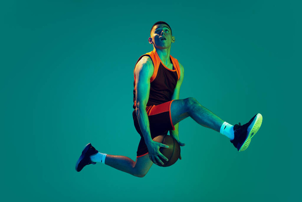 Portrait of young man, professional backetball player in motion, training isolated over blue background in neon light. Jump shot. Concept of sport, team game, action, active lifestyle, ad - Foto, Imagen