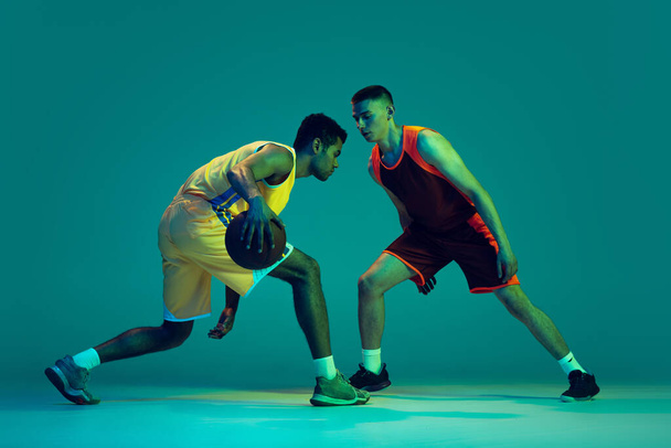 Porträt zweier Männer, Basketballprofis in Bewegung, die isoliert über blauem Studiohintergrund in Neonlicht trainieren. Dribbeln. Konzept von Sport, Teamspiel, Action, aktivem Lebensstil, Werbung - Foto, Bild