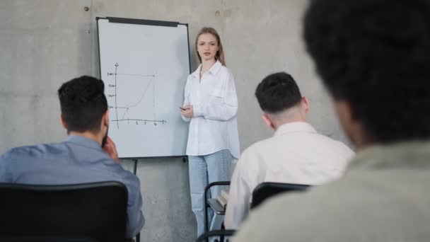 Young female teacher stands near flip chart in classroom at lesson explains work plan tells new topic caucasian girl tutor asks question diverse students raise hands correct answers business training - Footage, Video